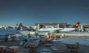 Samolot Lufthansa na płycie lotniska, gotowy do przyjęcia bagażu podręcznego. W tle budynek terminala i inne samoloty.