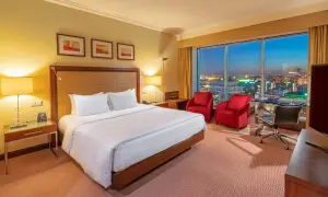 Przytulny pokój w hotelu Hilton Warszawa z widokiem na nocne miasto. Duże łóżko, dwa czerwone fotele i biurko.