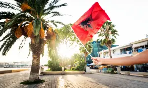Dłoń trzyma flagę Albanii na tle palmy i słońca. Czy Albania jest w Schengen? To pytanie nurtuje wielu podróżnych.