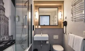 Łazienka w hotelu Campanile Warszawa z prysznicem, umywalką, toaletą i suszarką do włosów. Na ścianie grafika Pałacu Kultury.