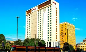 Hampton by Hilton Warsaw, nowoczesny hotel z charakterystycznym logo, góruje nad miejskim krajobrazem. Obok niego żółty budynek i tramwaj w ruchu.
