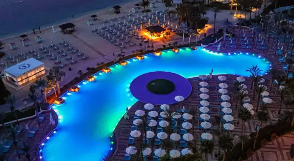 Nocny widok na luksusowy basen i plażę w Dubaju. Błękitna woda basenu, oświetlona kolorowymi światłami, otoczona leżakami i palmami.