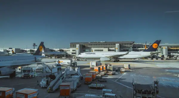 Samolot Lufthansa na płycie lotniska, gotowy do przyjęcia bagażu podręcznego. W tle budynek terminala i inne samoloty.