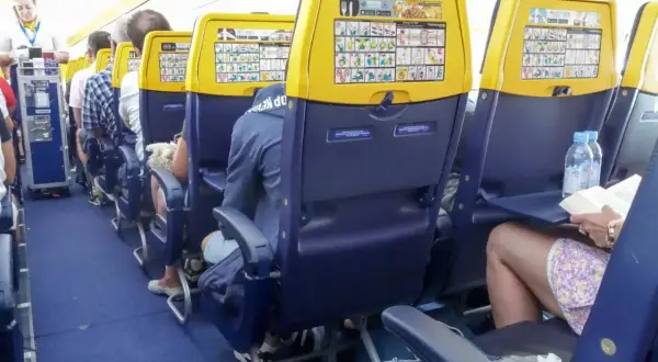 Wnętrze samolotu Boeing 737 Ryanair. Pasażerowie siedzą w rzędach, na oparciach foteli widoczne są instrukcje bezpieczeństwa.