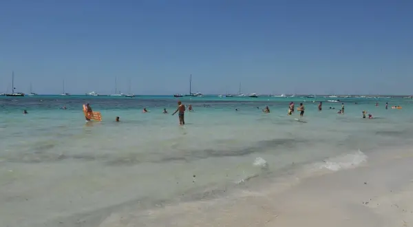 Słoneczny dzień na plaży Es Trenc. Ludzie kąpią się w turkusowej wodzie, a na horyzoncie widać żaglówki.