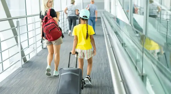 Młoda kobieta z plecakiem i dziewczynka z walizką idą korytarzem lotniska. To może być ich pierwszy wyjazd nieletniego za granicę bez rodziców.