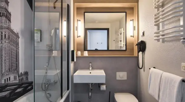 Łazienka w hotelu Campanile Warszawa z prysznicem, umywalką, toaletą i suszarką do włosów. Na ścianie grafika Pałacu Kultury.