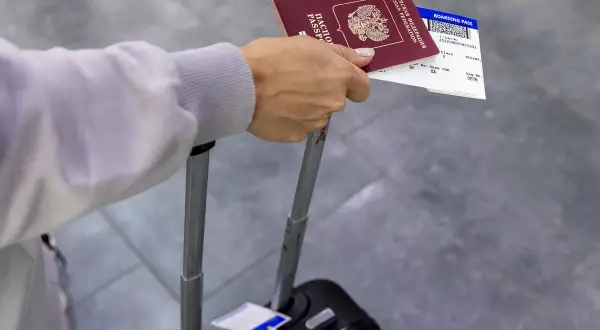 Dłoń trzyma paszport i kartę pokładową, która jest biletem na samolot. Obok walizka.