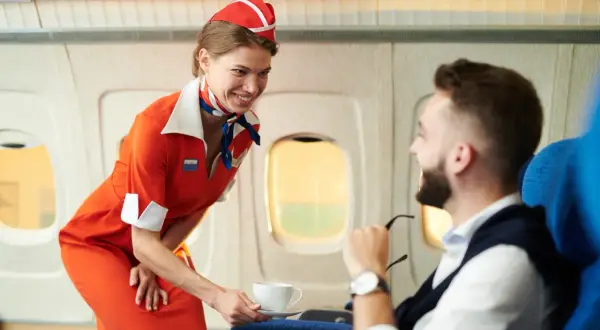 Uśmiechnięty stewardesa w pomarańczowym stroju podaje kawę pasażerowi w samolocie.