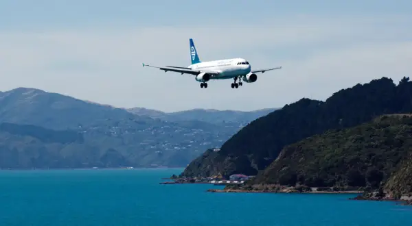 Samolot Air New Zealand ląduje nad turkusową wodą, zbliżając się do wybrzeża. Zastanawiasz się, ile kosztuje lot do Nowej Zelandii?
