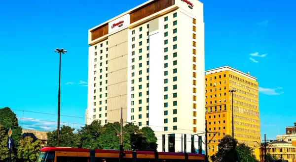 Hampton by Hilton Warsaw, nowoczesny hotel z charakterystycznym logo, góruje nad miejskim krajobrazem. Obok niego żółty budynek i tramwaj w ruchu.