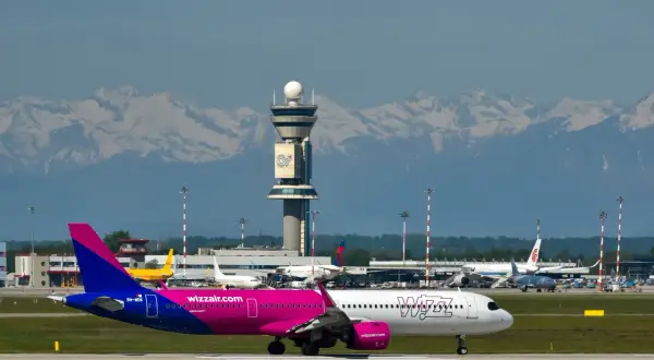 Różowy samolot Wizz Air na płycie lotniska, z wieżą kontroli lotów i ośnieżonymi górami w tle. Jak wydrukować bilety Wizzair?