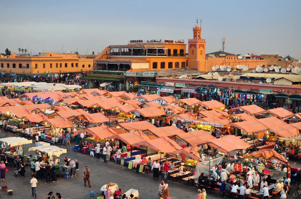 Targowisko w Marrakeszu, gdzie można znaleźć wiele produktów i poznać ceny. W tle budynki i minaret.