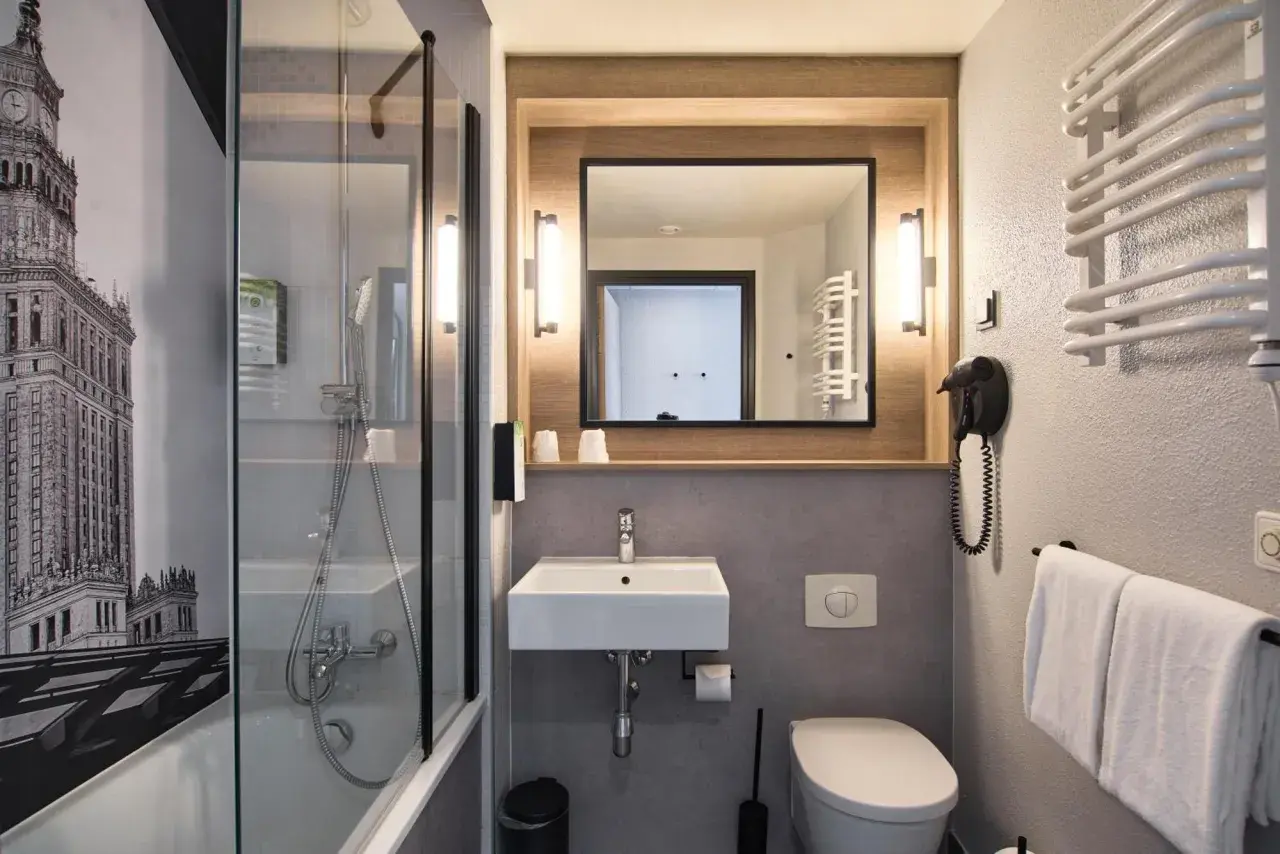 Łazienka w hotelu Campanile Warszawa z prysznicem, umywalką, toaletą i suszarką do włosów. Na ścianie grafika Pałacu Kultury.