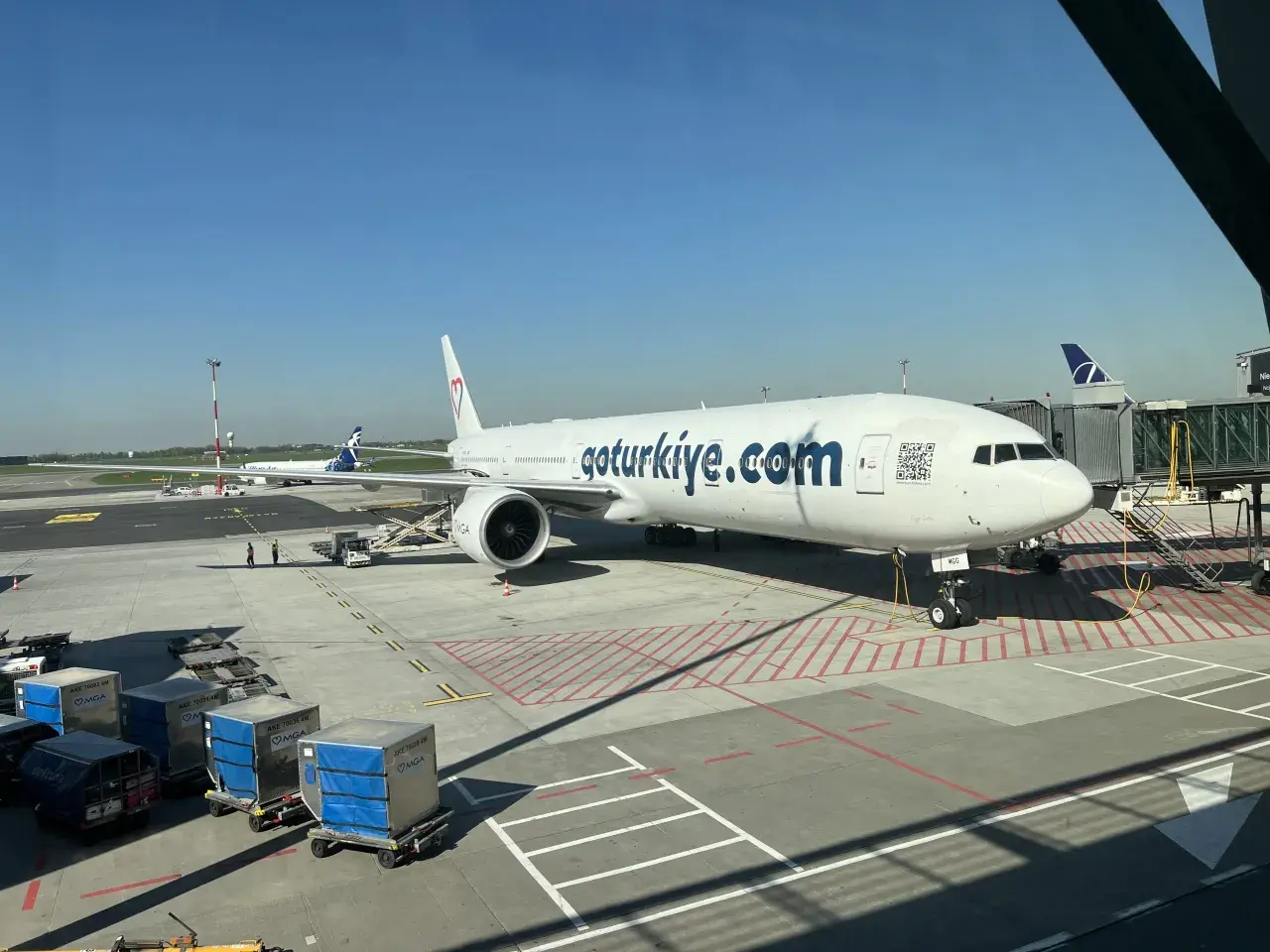 Samolot linii lotniczych MGA z napisem "goturkiye.com" na lotnisku. W tle widać inny samolot i rękaw.