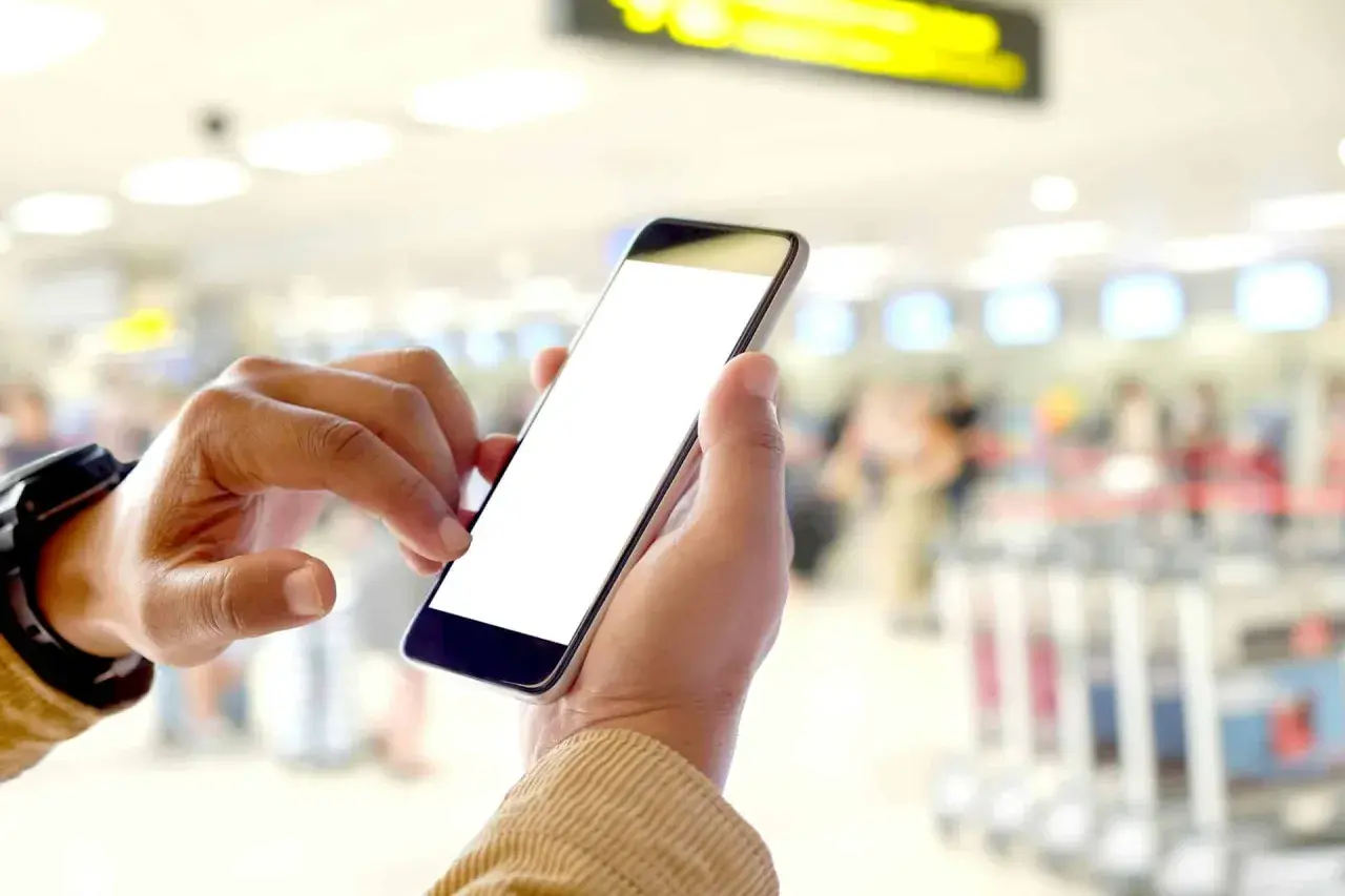 Ręce trzymają smartfon z pustym ekranem. W tle widać zamazany terminal lotniczy, sugerujący, że to idealny moment na **lot odprawa online kiedy** tylko chcesz.