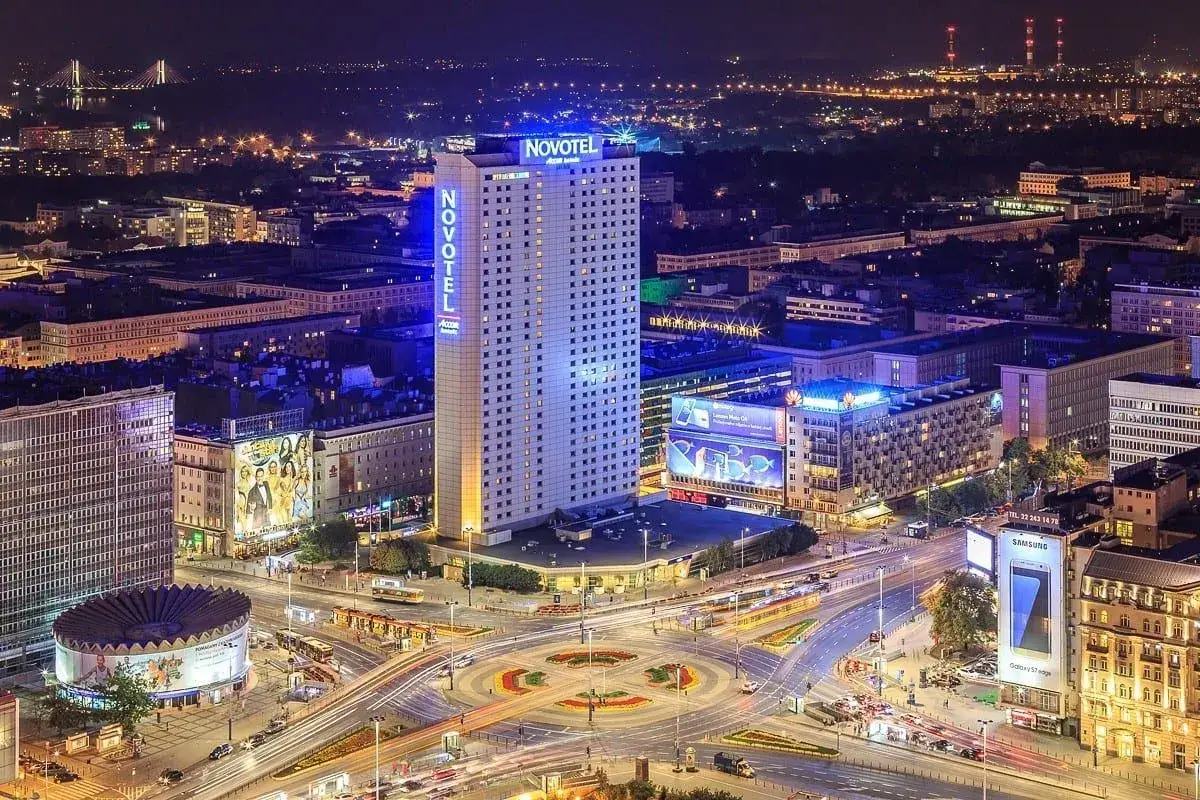Nocny widok na centrum Warszawy z oświetlonym Novotelem. Widać ruch uliczny i inne budynki.