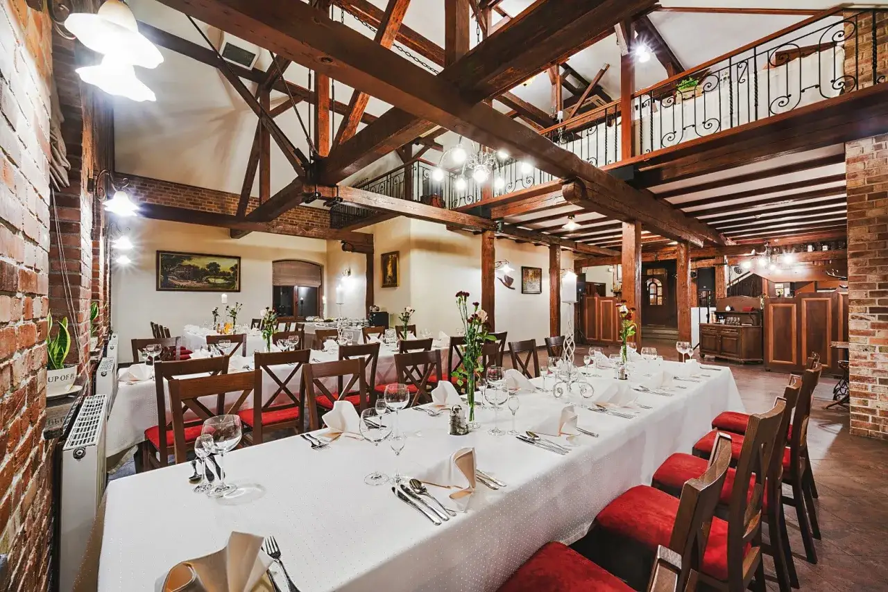 Elegancka sala bankietowa w hotelu Młyn Elbląg, z drewnianymi belkami stropowymi i ceglanymi ścianami, gotowa na przyjęcie gości.