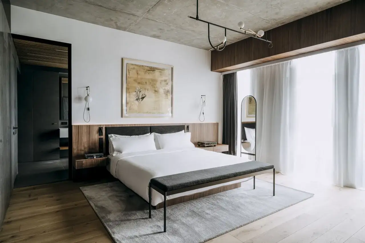 Elegancka sypialnia w hotelu Nobu Warszawa z białym łóżkiem, szarą ławką i nowoczesnym oświetleniem.