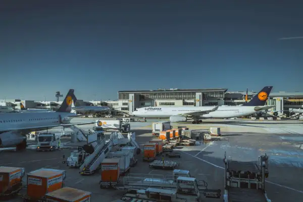 Samolot Lufthansa na płycie lotniska, gotowy do przyjęcia bagażu podręcznego. W tle budynek terminala i inne samoloty.