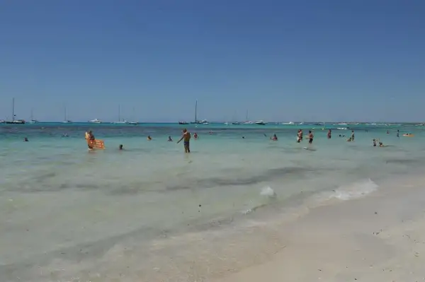 Słoneczny dzień na plaży Es Trenc. Ludzie kąpią się w turkusowej wodzie, a na horyzoncie widać żaglówki.