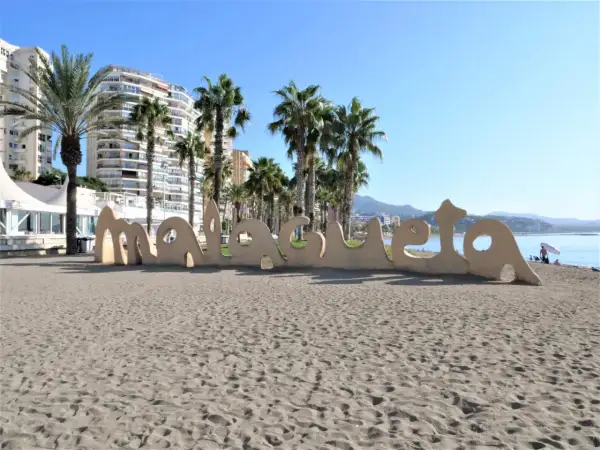 Malaga plaże: piasek, palmy i napis "Malaga" na tle budynków i morza.
