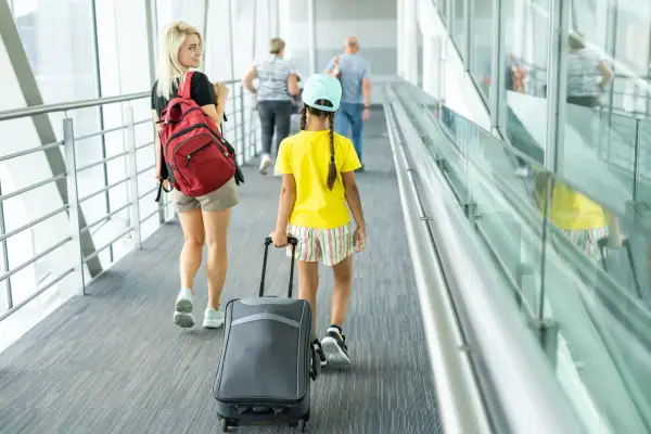 Młoda kobieta z plecakiem i dziewczynka z walizką idą korytarzem lotniska. To może być ich pierwszy wyjazd nieletniego za granicę bez rodziców.