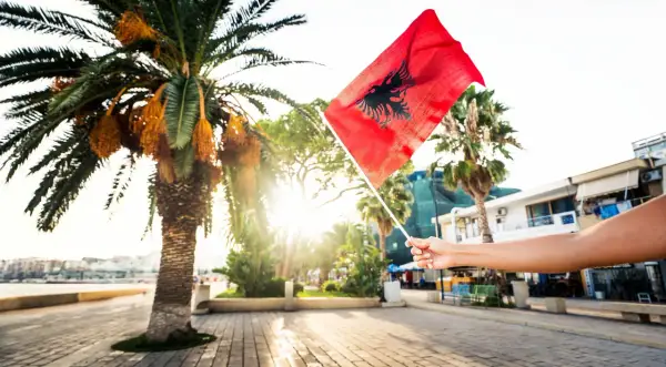 Dłoń trzyma flagę Albanii na tle palmy i słońca. Czy Albania jest w Schengen? To pytanie nurtuje wielu podróżnych.