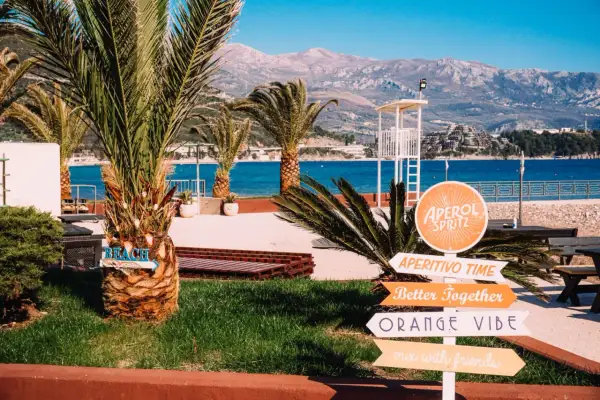 Palmy, morze i góry w Czarnogórze. Znaki wskazują plażę, Aperol Spritz i "Orange Vibe". Idealne miejsce na wakacje.