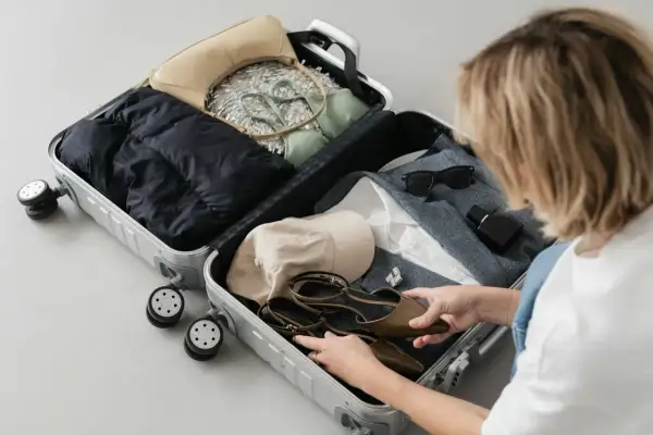Kobieta pakuje do walizki: kurtkę, torebkę, bieliznę, czapkę, buty, okulary i perfumy. Gotowa na podróż, pakuje swój enter air bagaż.