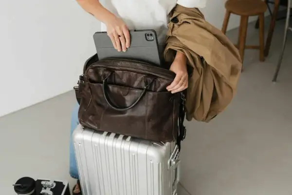 Kobieta pakuje tablet do brązowej skórzanej torby, gotowa na podróż. Jej bagaż, w tym walizka, czeka na szybki transport.