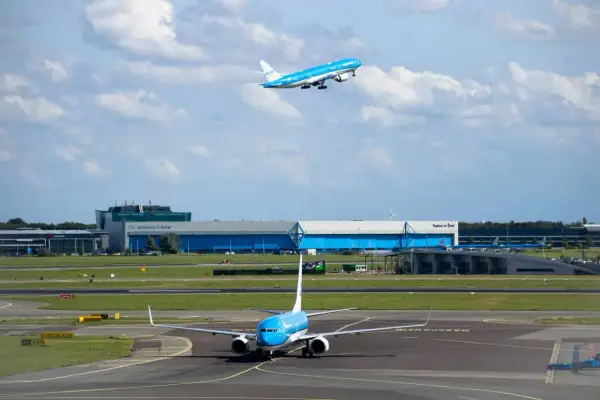 Dwa samoloty linii KLM na płycie lotniska i w powietrzu. Jeden kołuje, drugi wznosi się na tle chmur i budynków lotniska.